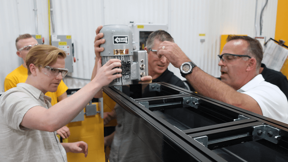Four people wearing safety glasses are assembling or inspecting a mechanical device in an industrial setting, emphasizing quality. One person holds a motor, while another points at components. Yellow walls and electrical equipment ensure a focus on safety in the background.