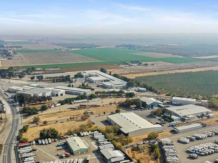An aerial view showcases a rural industrial area, featuring Dynatect's warehouses and buildings amid sprawling farmland. Fields stretch into the distance, while rows of parked vehicles, including trailers, hint at bustling careers within. The sky is clear with some cloud cover.