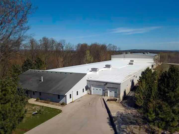 A large industrial building with a white roof and several entrances is surrounded by trees, likely housing Dynatect operations. The structure sits in an open area beneath a clear blue sky. A driveway leads to a loading dock at the side, possibly where new careers in logistics are born.