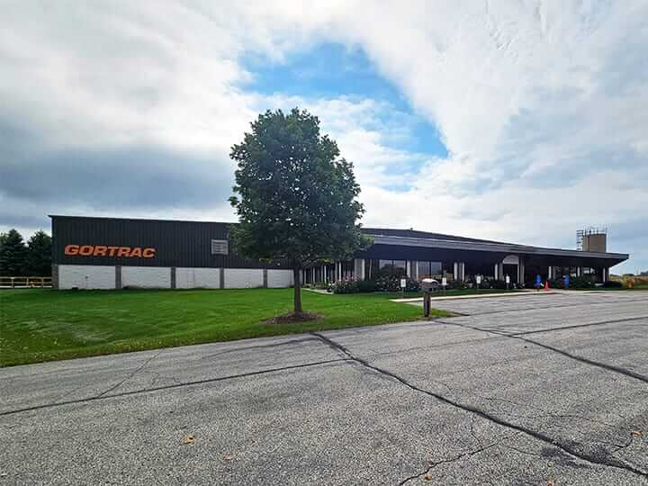 A large industrial building with the word GORTRAC on the side is visible, hinting at exciting Dynatect careers within. In front of the building is a neatly trimmed lawn with a solitary tree under a partly cloudy sky, while the foreground reveals a broad paved area promising job opportunities.